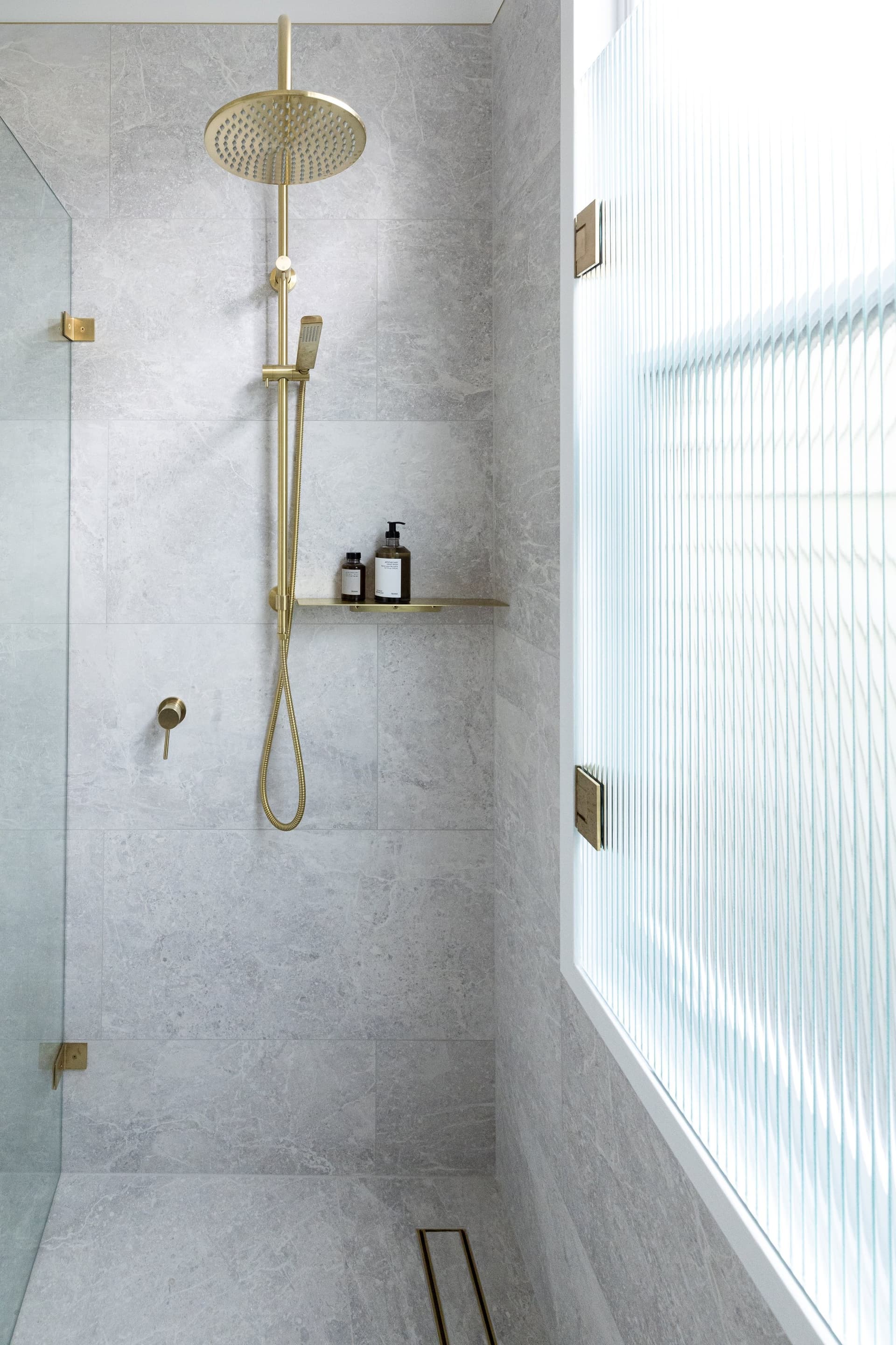 Stone-tiled shower with brass rainhead beside fluted glass